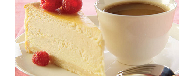 A slice of white chocolate cheesecake, topped with raspberries, with a cup of coffee.