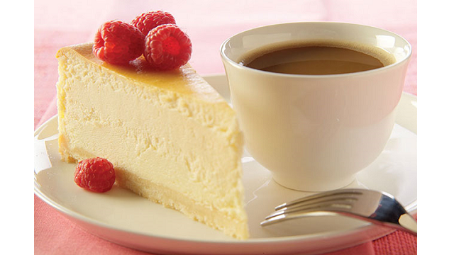 A slice of white chocolate cheesecake, topped with raspberries, with a cup of coffee.